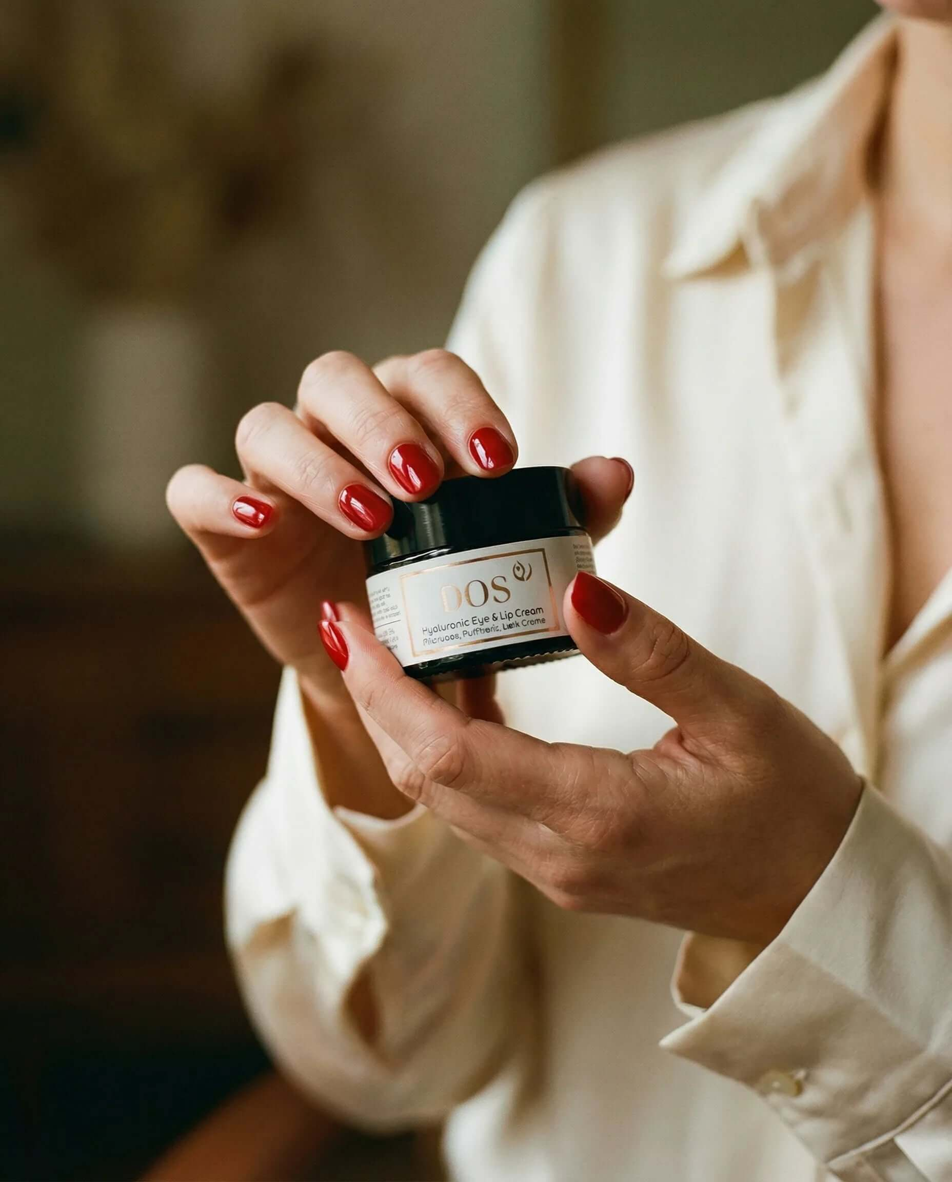 Hyaluronic eye cream in stylish jar held by a person with red nails, showcasing deep hydration for eyes and lips. DOS Cosmetics