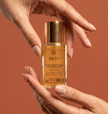 A bottle of DOS Oxygen Species Mandelicum night repair amine skin serum oil is held between two hands against a solid terracotta background. The product is a clear glass bottle filled with golden-yellow oil, topped with a metallic gold cap.